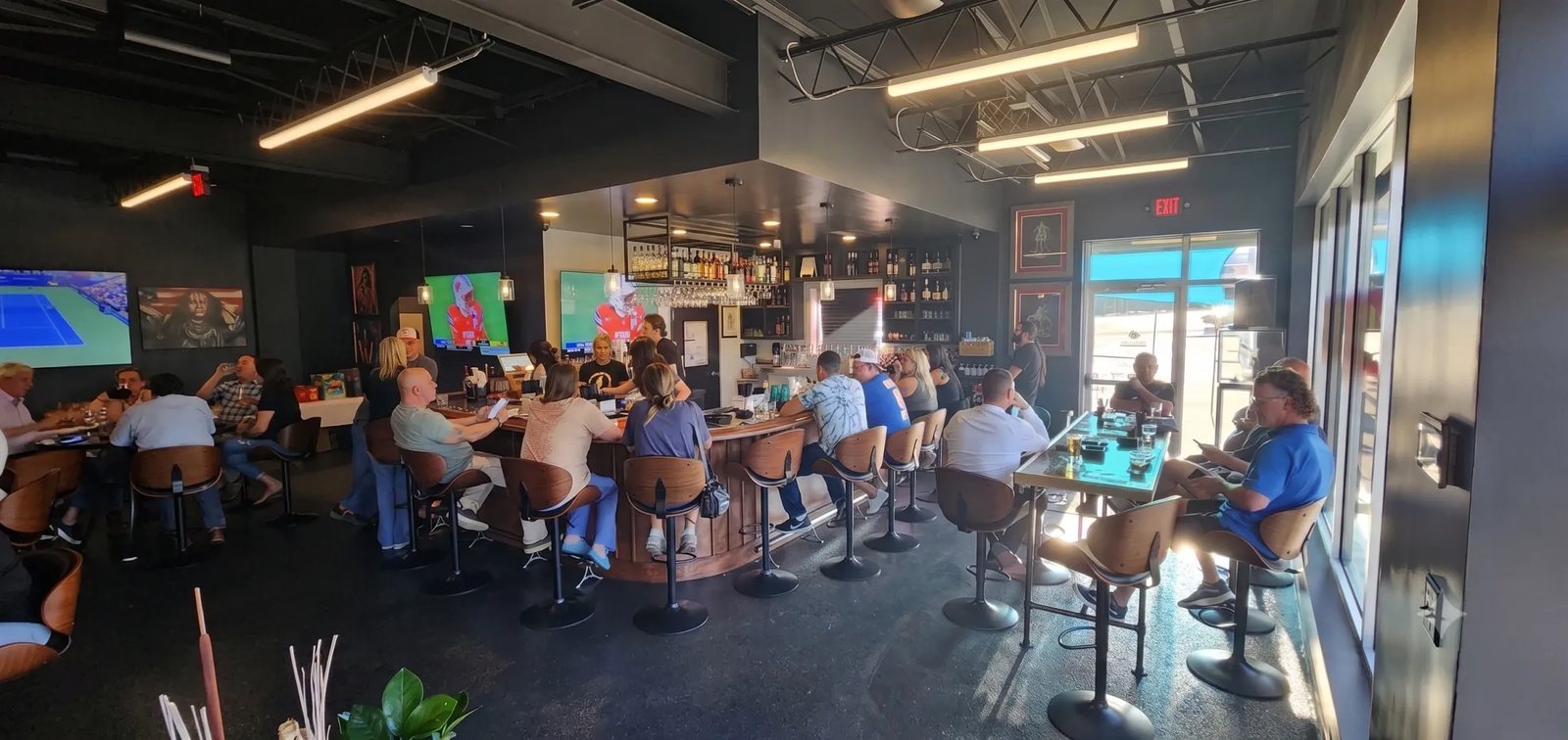 Guests at Palomino Cigar Lounge's curved bar and high-top tables, with TVs on the walls and daylight through the front windows