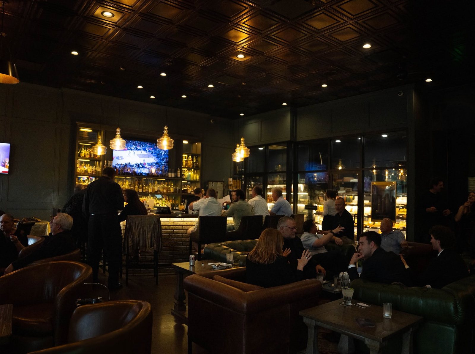 The Royale's main room at night with guests on leather sofas, the bar under glass pendant lights, and a lit glass humidor along the right wall